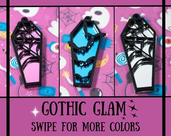 May include: Three black coffin-shaped charms with different colored mirrored inserts. The charms are pink, blue, and silver. The text "GOTHIC GLAM" and "SWIPE FOR MORE COLORS" is displayed below the charms.