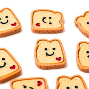 May include: Close-up of several small, smiling toast-shaped charms. Each charm has a yellow bread interior, a brown crust, a black smiley face, and a red heart. The charms are scattered on a white background.