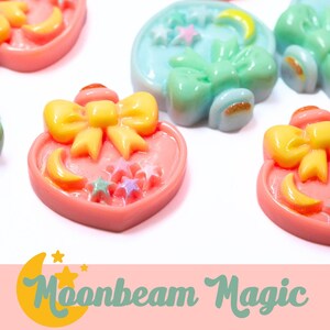 May include: A collection of heart-shaped resin charms in shades of pink, blue, and green, adorned with yellow bows, crescent moons, and stars. The charms have a glossy finish and are arranged on a white surface. The text "Moonbeam Magic" is visible.