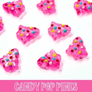 May include: Pink, translucent Christmas tree-shaped candies with colorful sprinkles. The candies are scattered on a white surface. The text "CANDY POP PINES" is displayed in pink at the bottom of the image.