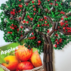 May include: A whimsical tree made of green pasta, red pasta, and brown licorice. The tree sits atop a book titled "Apples" with a basket of apples on the cover. The tree's leaves are made of green pasta, with red pasta representing the apples.