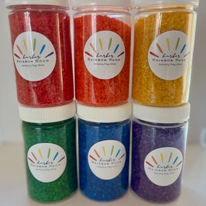 May include: Six clear plastic jars filled with colorful sensory rice. The jars are arranged in two rows of three, with red, orange, and yellow rice on top, and green, blue, and purple rice below. Each jar has a white lid and a label that reads "harbor RAINBOW ROOM Sensory Play Shop."