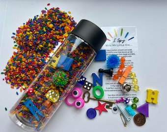 May include: A clear plastic sensory bottle filled with colorful rice and small toys. The bottle is next to a pile of matching rice and various objects, including dice, buttons, and a key. A card reads "I Spy".