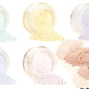 May include: Six small glass jars with lids, each containing a different colored powder. The powders are light green, yellow, light purple, white, pink, and beige. The jars are arranged in two rows of three, with the powders spilling out of the jars.