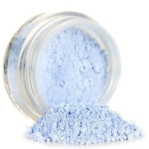 May include: A small glass jar filled with light blue loose powder. The powder is spilling out of the jar, forming a small pile on a white surface.