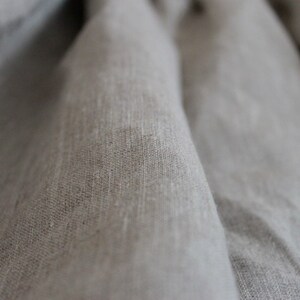 May include: Close-up of a light gray linen fabric with a textured weave. The fabric is folded and draped, creating soft folds and shadows.