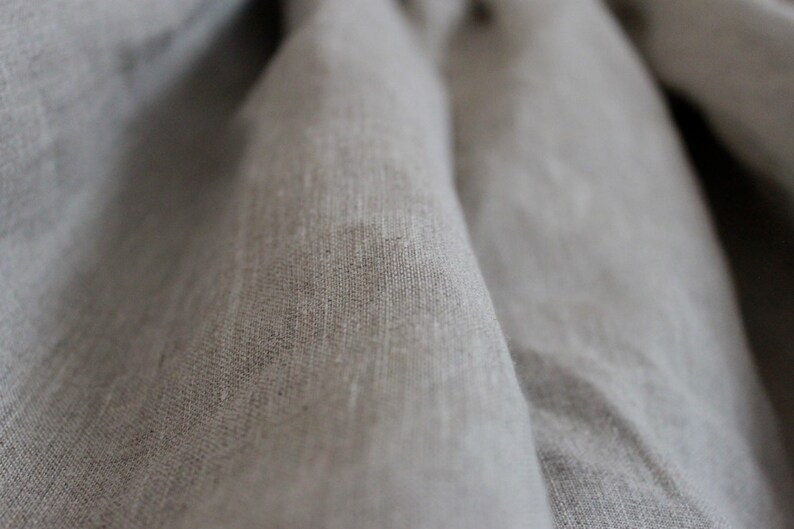 May include: Close-up of a light gray linen fabric with a textured weave. The fabric is folded and draped, creating soft folds and shadows.