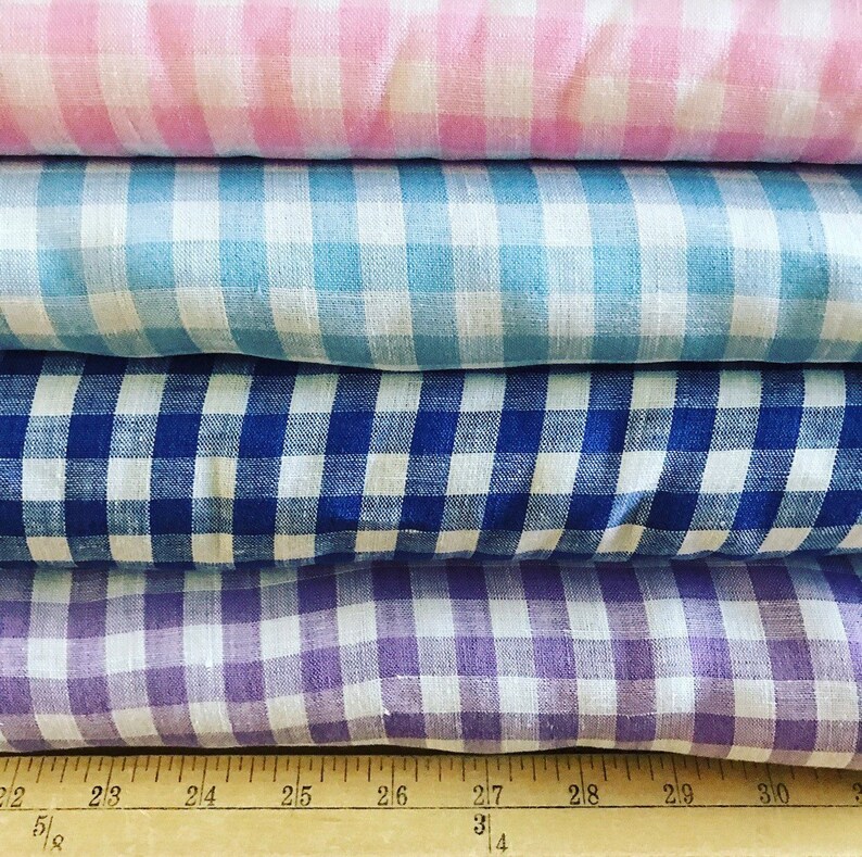 May include: A stack of four fabrics in pink, light blue, navy blue, and purple gingham patterns. The fabrics are folded and stacked on top of each other. A wooden ruler is at the bottom of the image.