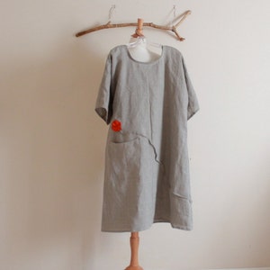 May include: A gray linen dress with a red flower detail and a single pocket. The dress is hanging on a wooden branch.
