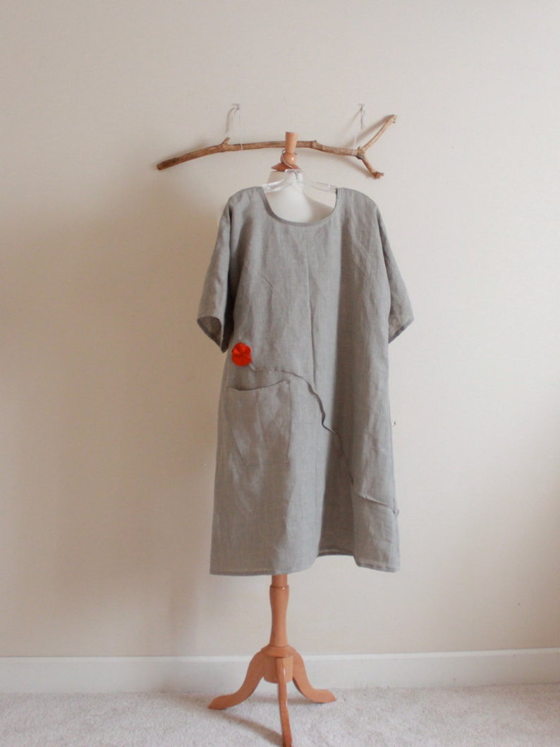 May include: A gray linen dress with a red flower detail and a single pocket. The dress is hanging on a wooden branch.