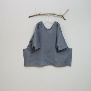 May include: A gray, loose-fitting tunic top with a wide neckline and short sleeves. The tunic is hanging on a clear hanger suspended from a small branch attached to the wall. The fabric appears to be a soft, woven material.