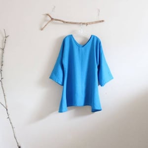 May include: A light blue linen top with a v-neck and 3/4 sleeves. The top is hanging on a wooden hanger against a white wall.