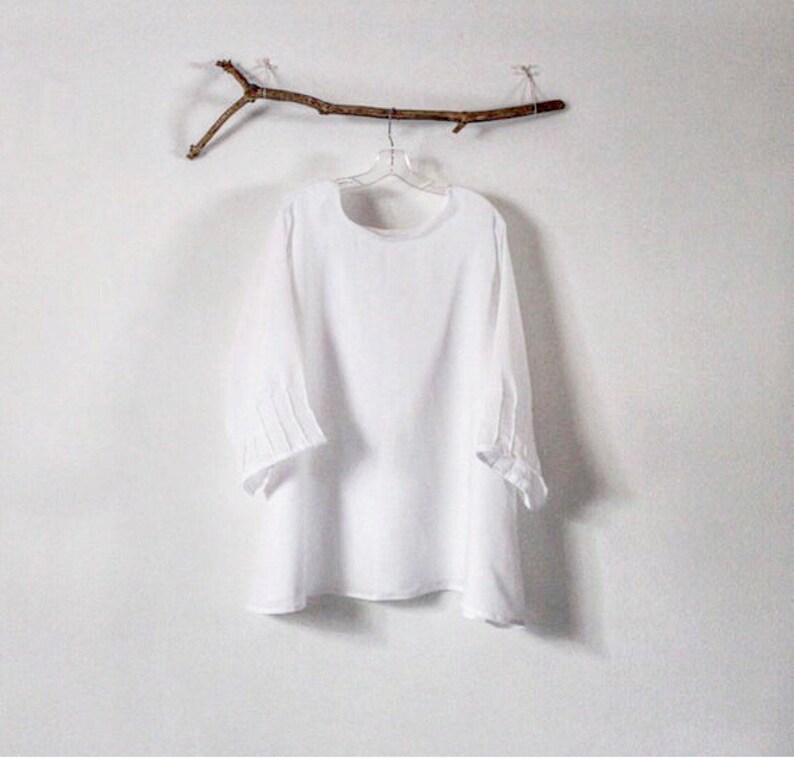 May include: A white, long-sleeved blouse with a round neckline and three-quarter sleeves. The sleeves have pleated details at the elbow. The blouse hangs on a clear hanger suspended from a decorative twig against a white wall.