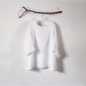 May include: A white, long-sleeved blouse with a round neckline and three-quarter sleeves. The sleeves have pleated details at the elbow. The blouse hangs on a clear hanger suspended from a decorative twig against a white wall.