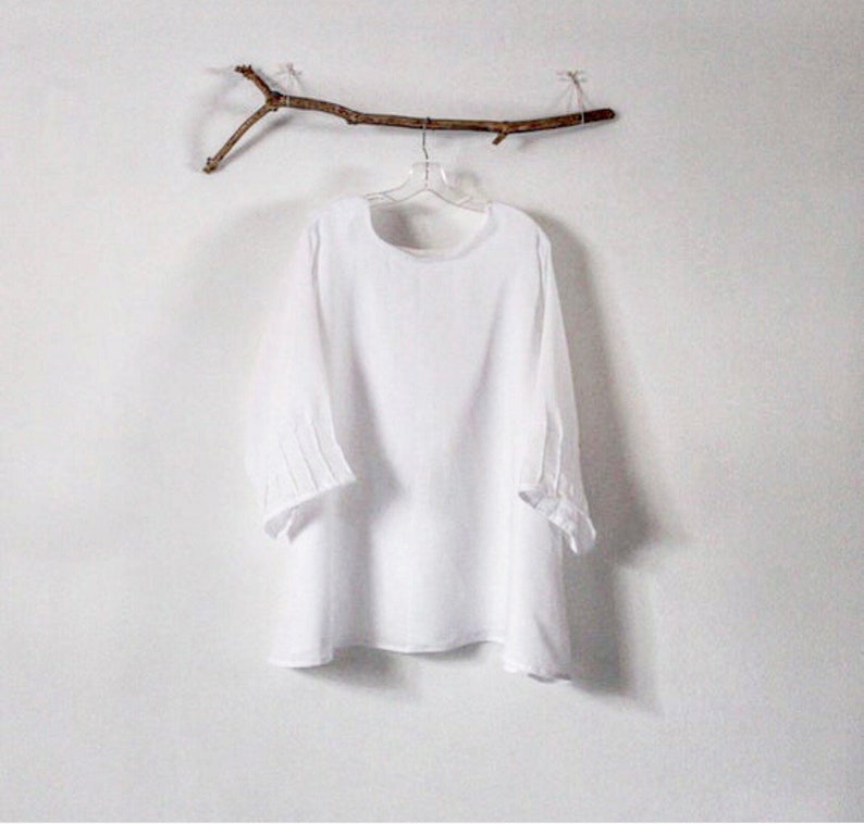 May include: A white, long-sleeved blouse with a round neckline and three-quarter sleeves. The sleeves have pleated details at the elbow. The blouse hangs on a clear hanger suspended from a decorative twig against a white wall.