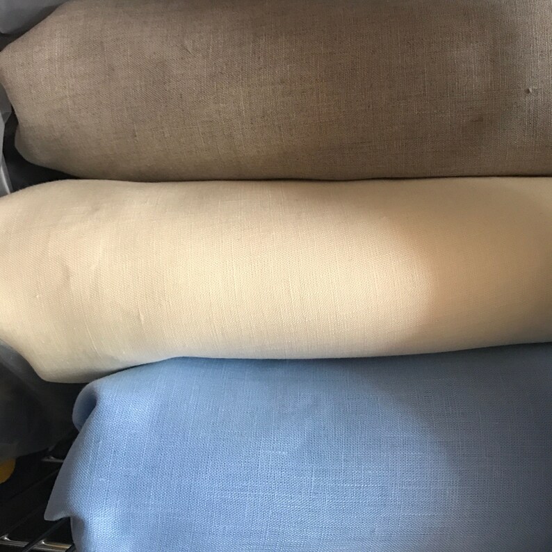 May include: Three rolled fabric pieces in neutral tones. The top piece is a light brown, the middle is a creamy off-white, and the bottom is a soft blue. The fabric appears to be linen or a similar woven material.