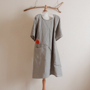 May include: A gray linen dress with a red flower detail and a single pocket. The dress is on a wooden mannequin stand.