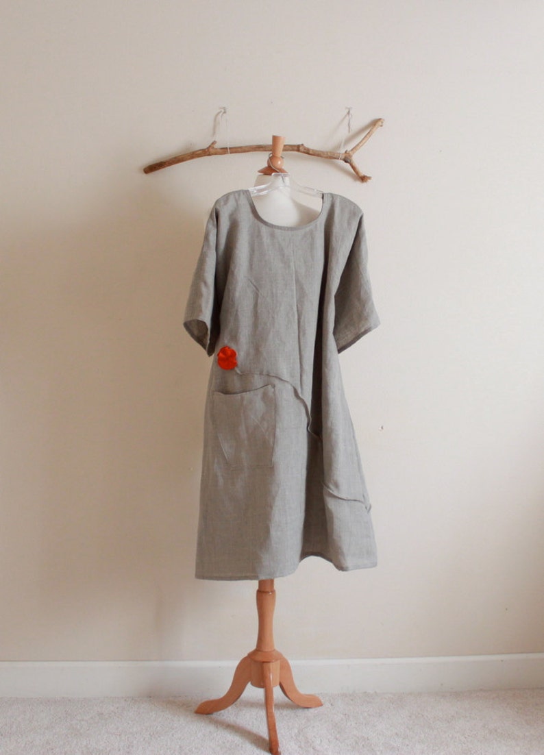 May include: A gray linen dress with a red flower detail and a single pocket. The dress is on a wooden mannequin stand.