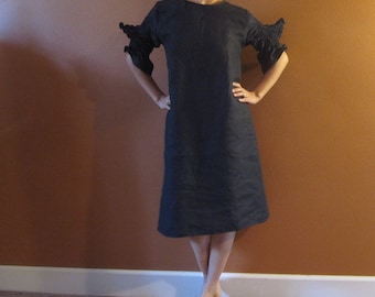 May include: A woman wearing a black linen dress with ruffled sleeves. The dress has a simple, elegant design and is knee-length.