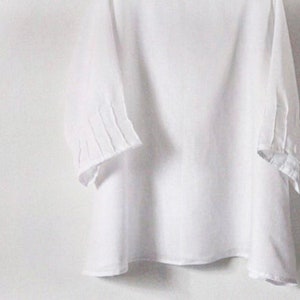May include: A white, three-quarter sleeve top with pleated details on the sleeves. The top is made of a lightweight fabric and has a simple, elegant design. The sleeves are approximately 15 inches long.