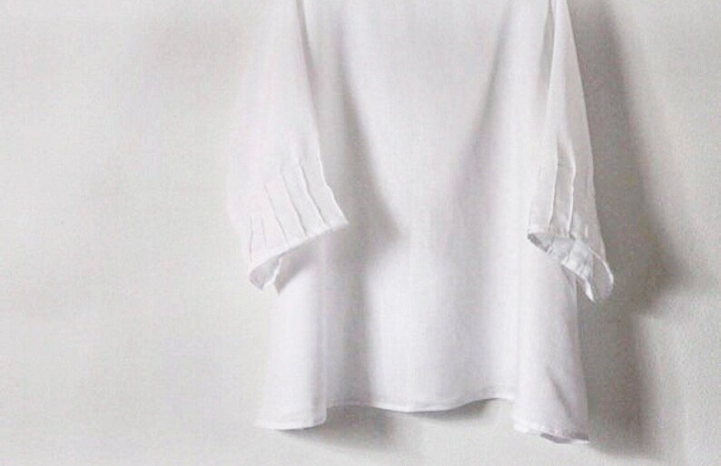 May include: A white, three-quarter sleeve top with pleated details on the sleeves. The top is made of a lightweight fabric and has a simple, elegant design. The sleeves are approximately 15 inches long.