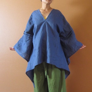 May include: A blue linen tunic with a V-neckline and long, wide sleeves. The tunic is asymmetrical, with a longer back hem. The model is wearing the tunic with green pants.