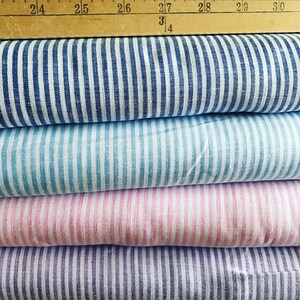 May include: A stack of striped fabric in shades of blue, light blue, pink, and purple. The fabric has vertical white stripes. A wooden ruler is placed on top of the fabric.