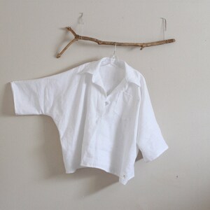 Pode incluir: Uma camisa branca de linho com um caimento solto e gola de botões. A camisa está pendurada em um galho de madeira.
