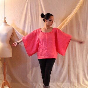May include: A woman wearing a bright pink, loose-fitting linen top with a scoop neck and wide sleeves. She is wearing black pants and has her hair pulled back in a bun.