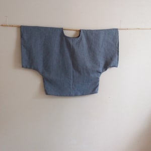 May include: A light blue linen top with a wide, loose fit and short sleeves. The top has a round neckline and is hanging on a wooden rod.