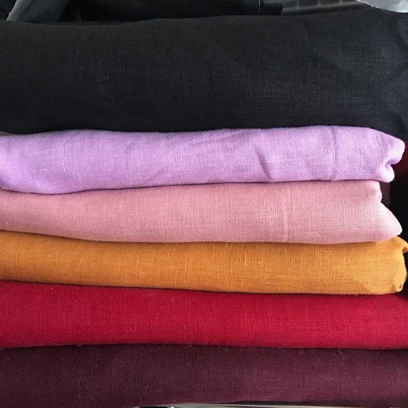 May include: A stack of folded linen fabric in various colors. The colors include black, lavender, light pink, mustard yellow, red, and burgundy. The fabric has a textured appearance.