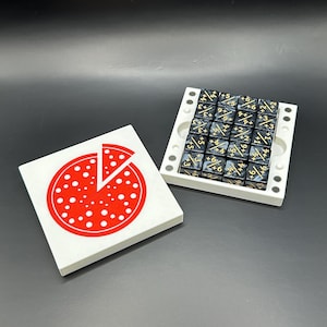 May include: A white square tile with a red pizza graphic and a missing slice sits next to a white tray holding 25 black dice with gold markings. The dice are arranged in a 5x5 grid. The pizza graphic has white dots.