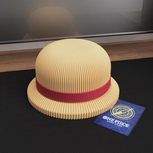 May include: A tan-colored hat with a red band, reminiscent of a straw hat, sits on a black surface. A blue One Piece card is next to the hat. The hat has a ribbed texture.