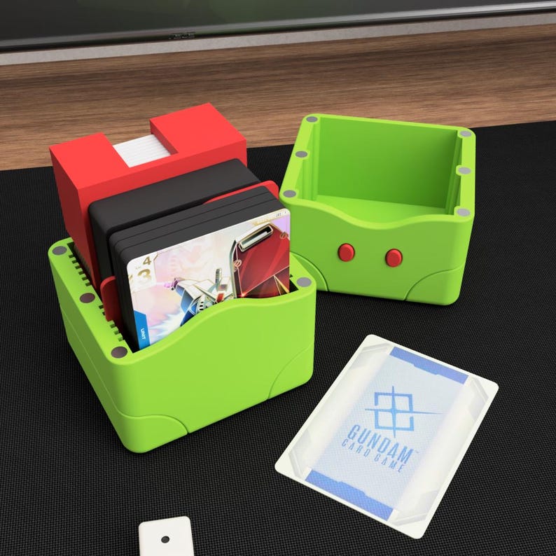 May include: A green card holder with a red top, holding a stack of cards. A second green card holder is next to it. A card with the text "GUNDAM CARD GAME" is in the foreground. A white die is also visible.