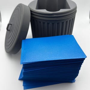 May include: A gray, 3D-printed miniature trash can with a removable lid, next to a stack of bright blue cards. The trash can has a ribbed design and a square opening. The lid has a handle.