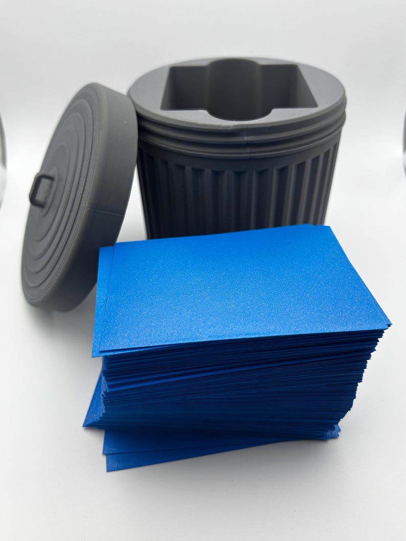 May include: A gray, 3D-printed miniature trash can with a removable lid, next to a stack of bright blue cards. The trash can has a ribbed design and a square opening. The lid has a handle.
