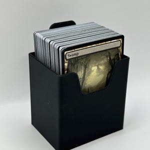 May include: A black card holder filled with a stack of trading cards. The top card is visible, displaying the word "Swamp" and a swampy landscape illustration. The holder is square with a curved cutout for easy card access.