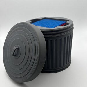 May include: A grey, 3D-printed container resembling a bin, with a removable lid. The container is open, revealing blue and red items inside. The lid is off to the side, showing the interior of the container.