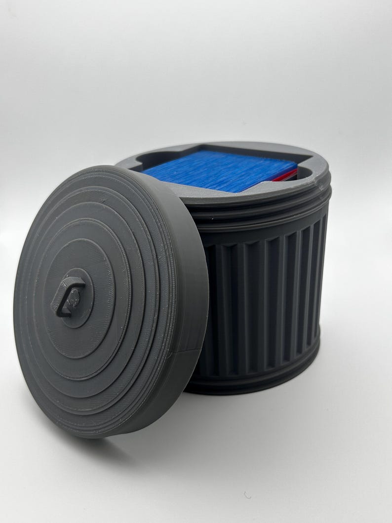 May include: A gray, 3D-printed container shaped like a trash can, with a removable lid. The container is open, revealing blue and red items inside. The lid is off to the side, showing the interior of the container.