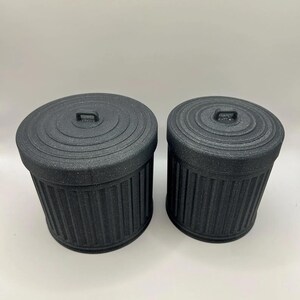 May include: Two black miniature trash cans with lids. The cans have a ribbed design and a small handle on the lid. The cans are made of a matte black material and are of different sizes.
