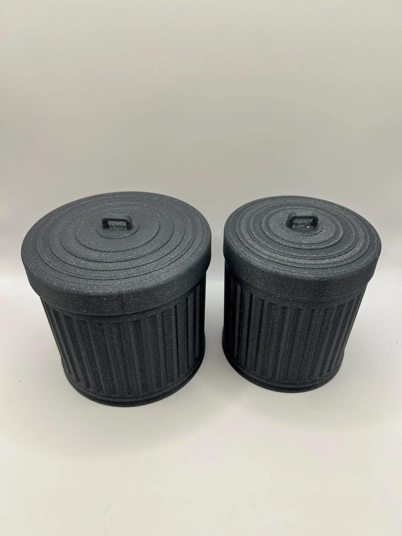 May include: Two black miniature trash cans with lids. The cans have a ribbed design and a small handle on the lid. The cans are made of a matte black material and are of different sizes.
