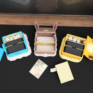 Pu&ograve; includere: Tre porta carte e dadi a tema personaggio in blu, marrone e giallo. Ogni contenitore ha un vano per carte e dadi. Sono visibili anche una carta collezionabile e un piccolo blocco note.