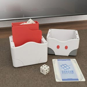 May include: White and red card game storage boxes with a dice and a card. The boxes have a wood grain texture. One box has red dividers. The card has the text "GUNDAM CARD GAME" and a blue and white design.