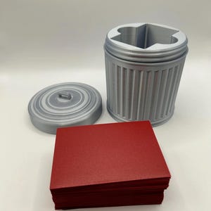 May include: A gray, 3D-printed trash can-shaped container with a lid and a stack of red cards. The container has a ridged design and a star-shaped opening. The lid is circular with a small handle.