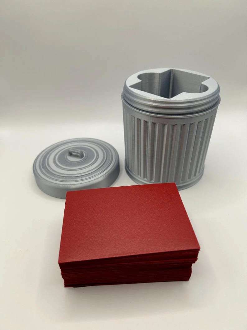 May include: A gray, 3D-printed trash can-shaped container with a lid and a stack of red cards. The container has a ridged design and a star-shaped opening. The lid is circular with a small handle.