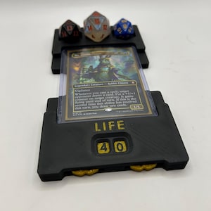 Könnte beinhalten: Schwarzer 3D-gedruckter Kartenhalter mit einer Magic: The Gathering-Karte, drei Würfeln und einem Lebenszähler. Die Karte befindet sich in einer Schutzhülle. Der Lebenszähler zeigt "LIFE 40" an. Die Würfel sind schwarz, transparent und blau mit weißen und goldenen Zahlen.