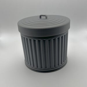 May include: A gray, miniature trash can with a matching lid. The can has vertical ribbed detailing and a small handle on the lid. The design is a solid, matte gray color.