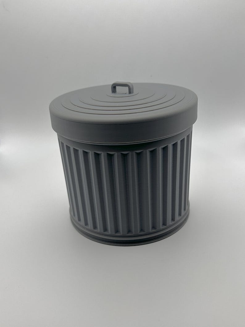 May include: A gray, miniature trash can with a matching lid. The can has vertical ribbed detailing and a small handle on the lid. The design is a solid, matte gray color.