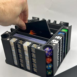 May include: A black, multi-compartment dice and card organizer. The organizer holds stacks of dice, cards, and other gaming accessories. A hand is placing a card into the top compartment. The organizer is made of black plastic.