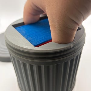 May include: A gray, 3D-printed trash can-shaped container with a lid. The container has vertical ridges and holds a stack of blue and red cards. A hand is reaching into the container.